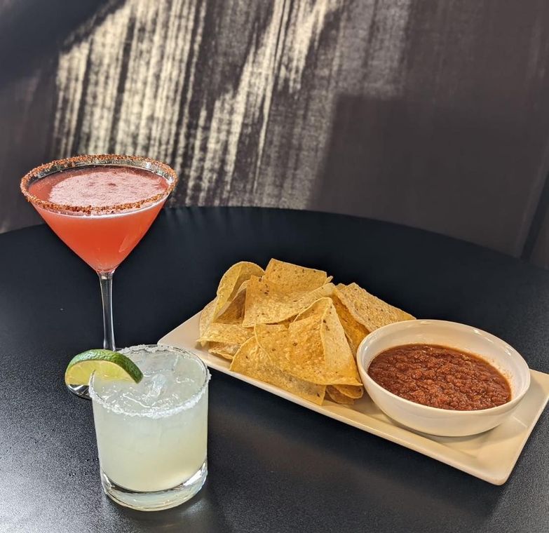 A martini and a margarita are on a table next to a plate of tortilla chips.