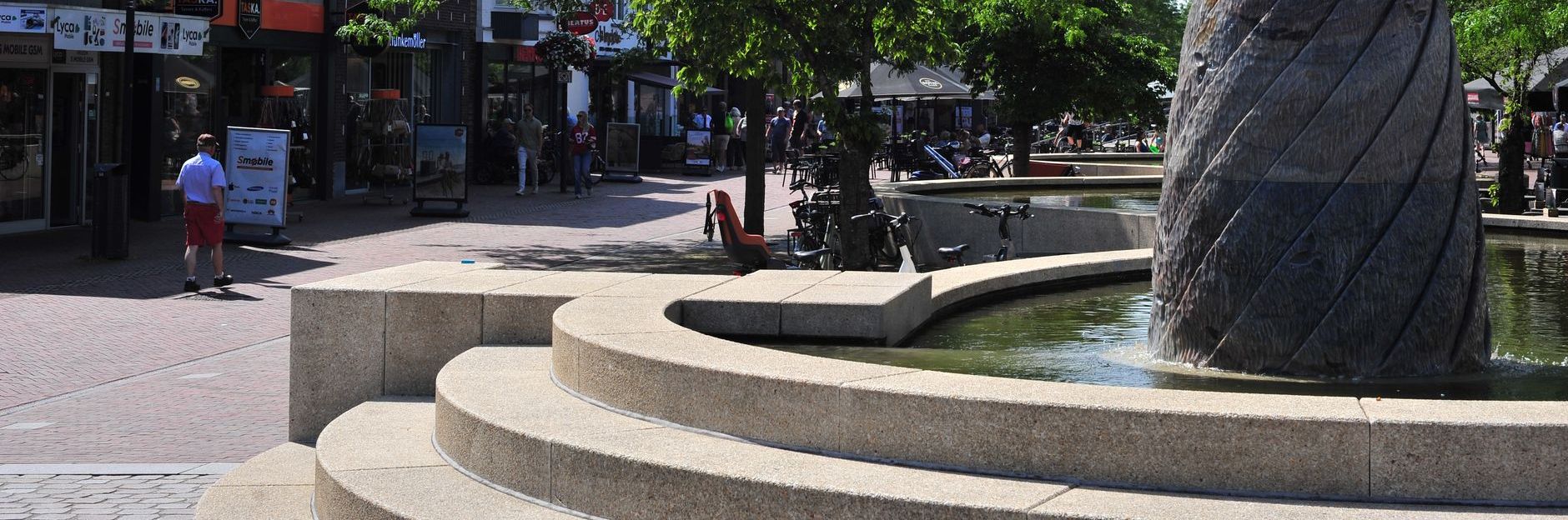 Een persoon loopt langs een stenen fontein op een plein met winkels en bomen op de achtergrond.