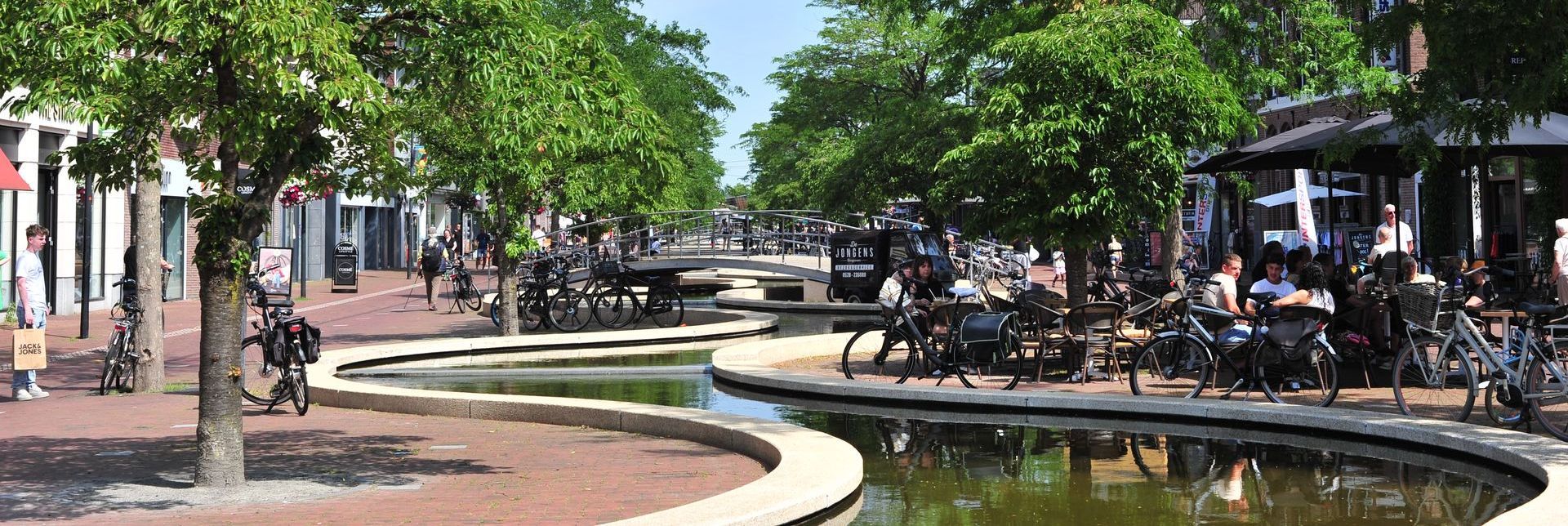 Een kanaal omzoomd met bomen en winkels, vol met fietsers en mensen te paard in een Europese stad.
