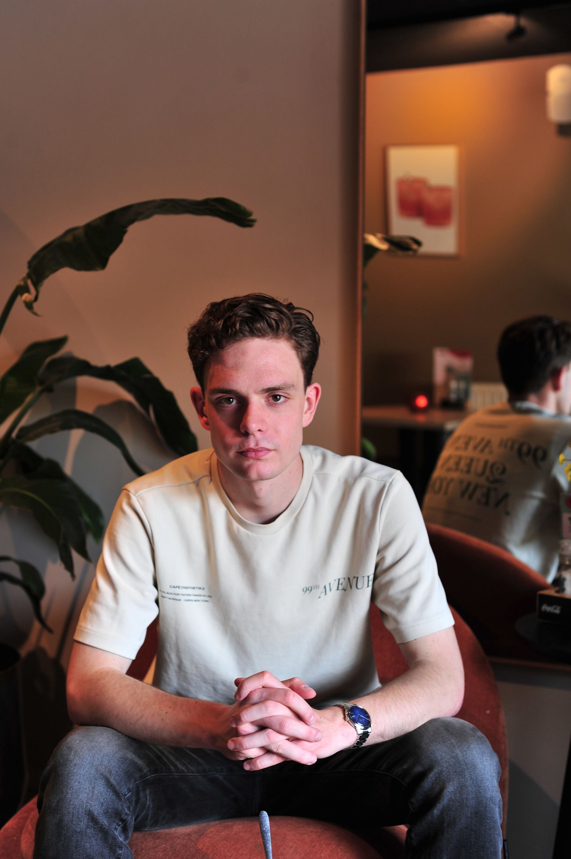 Een jongeman met krullend haar zit op een stoel, zijn handen gevouwen. Hij draagt een bruin T-shirt en een spijkerbroek. Een spiegel reflecteert zijn rug.