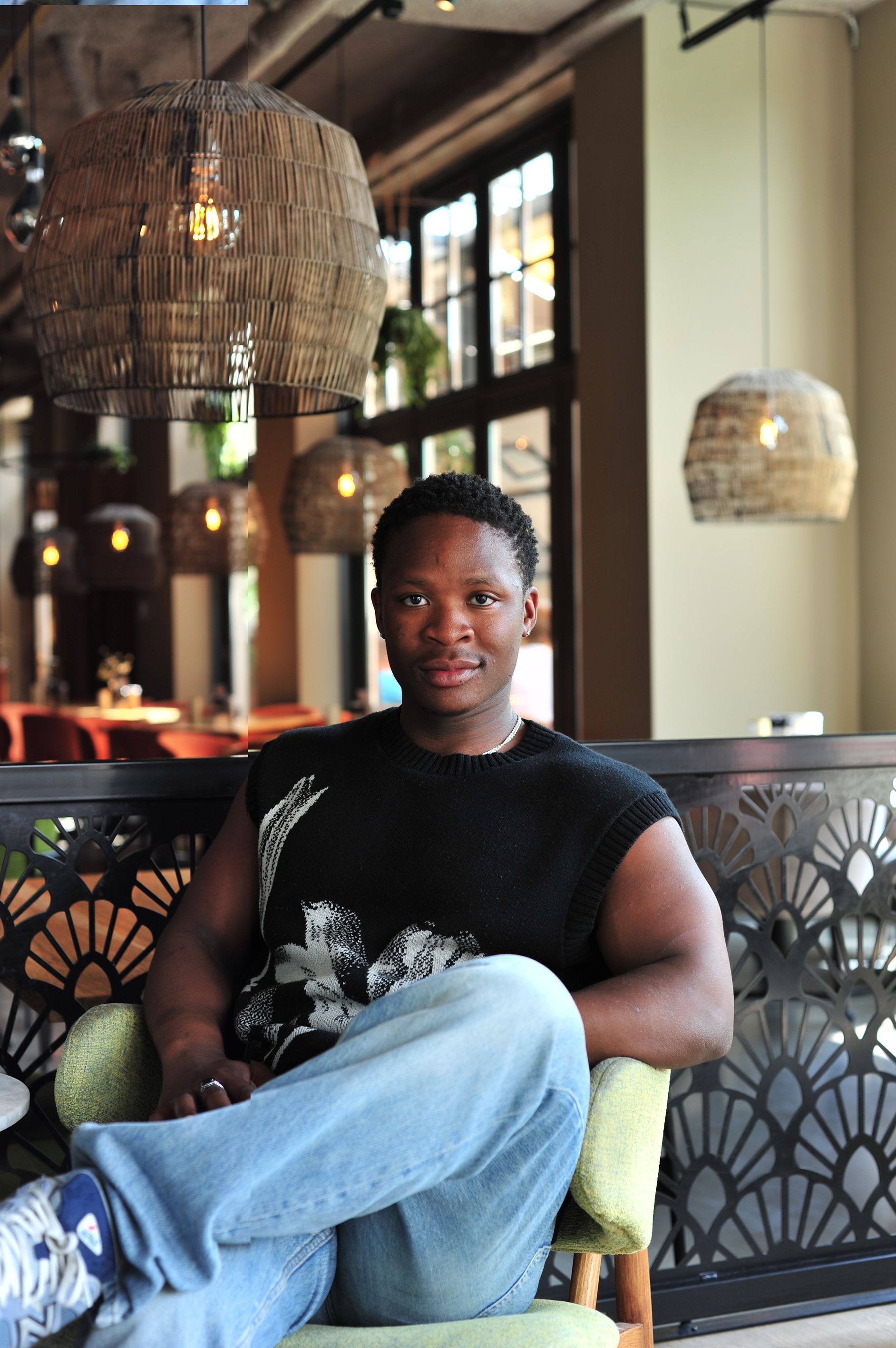 Een man in een zwart vest en spijkerbroek zit ontspannen in een stoel. Hij zit binnen bij een raam met decoratieve lampen.