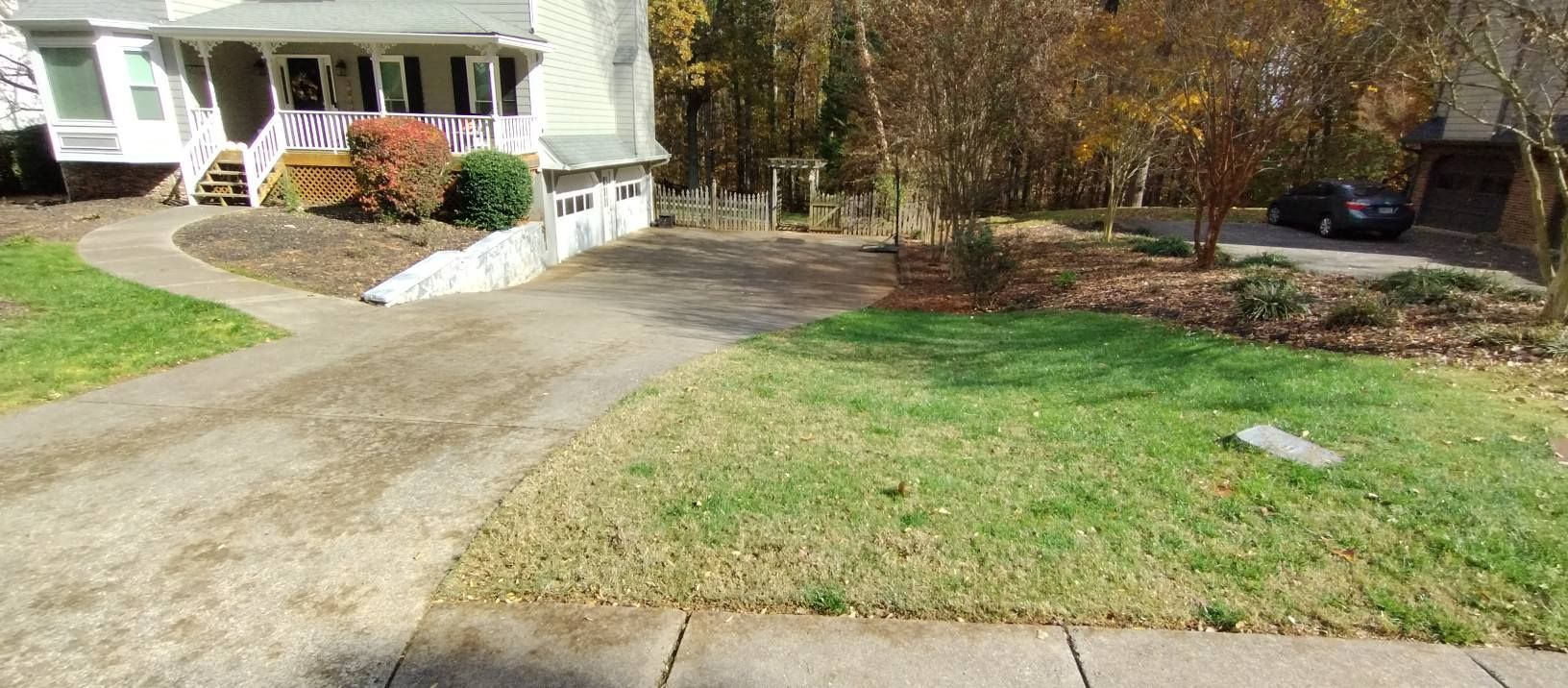 A paved driveway leads to a light-colored house with a small front yard and surrounding trees on a sunny day.