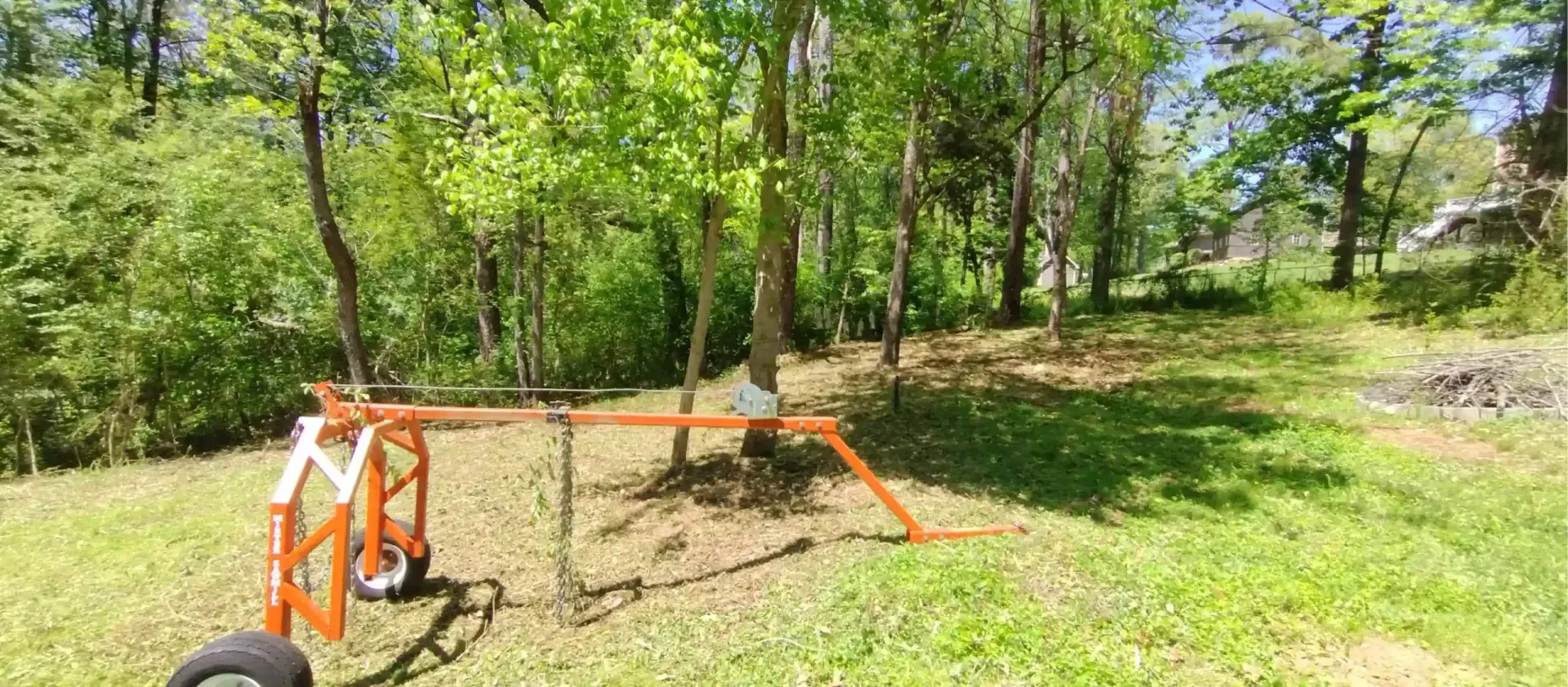 An orange, wheeled logging arch tool stands in a grassy, sunlit wooded area.