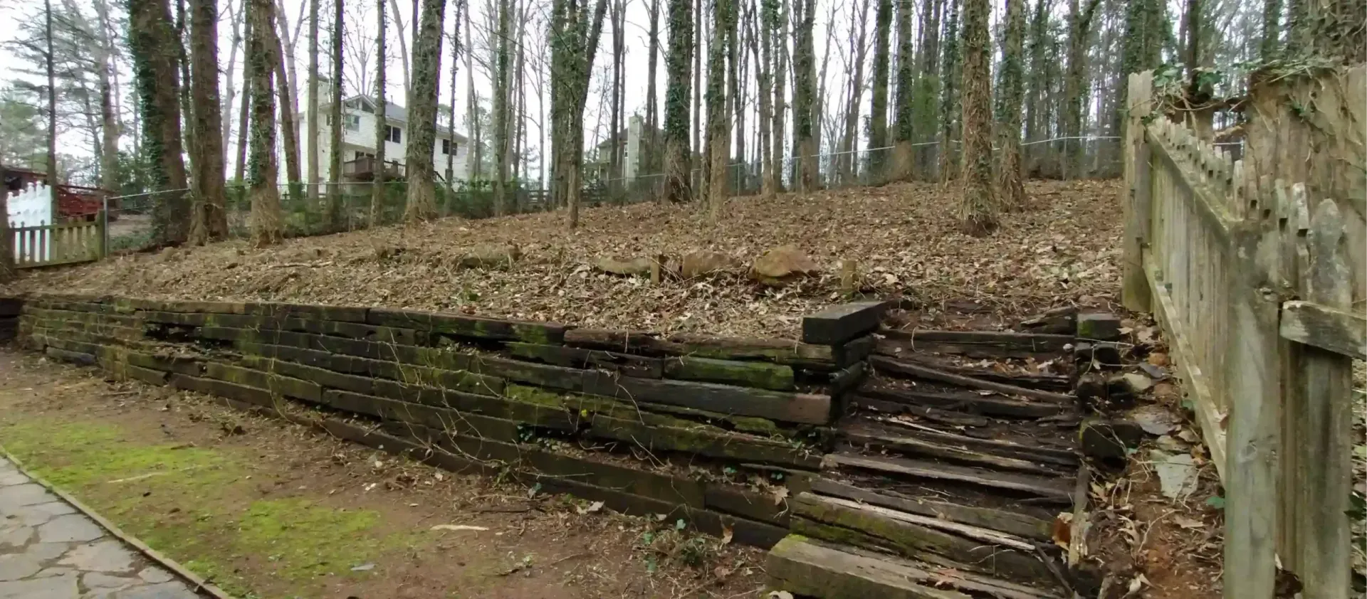 A weathered timber retaining wall lines a yard covered in fallen leaves, with wooden steps leading up a slight incline.