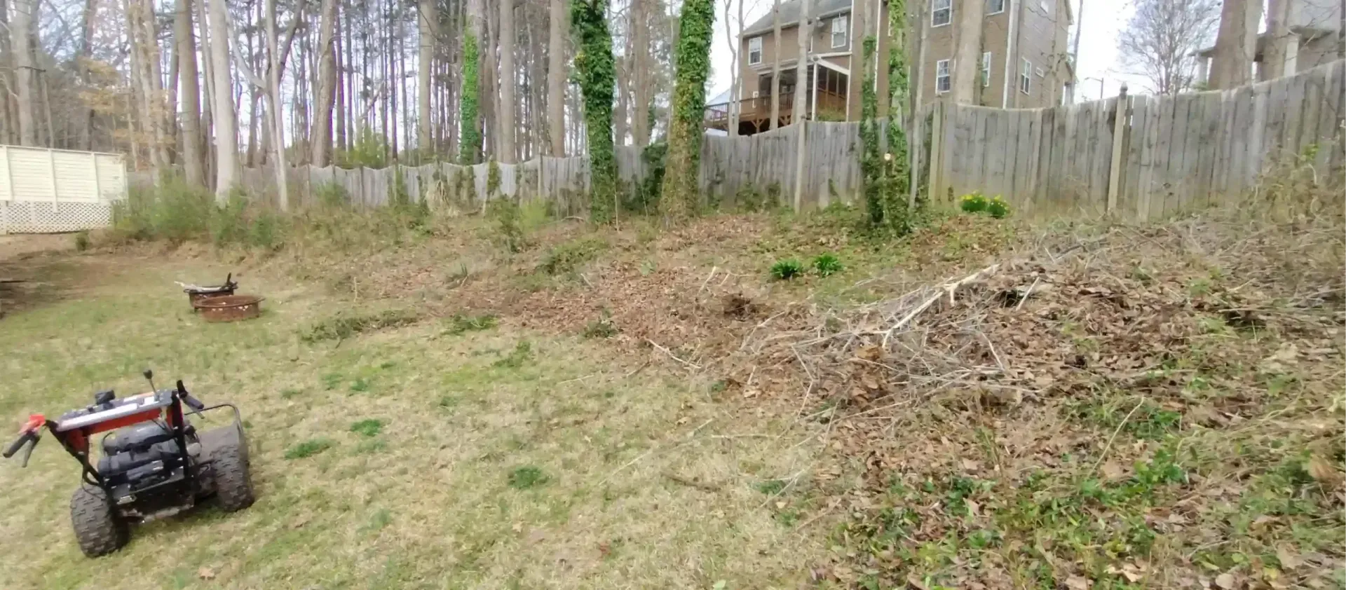 A brush mower sits on a grassy slope leading to a wooded area behind a fence in a backyard.