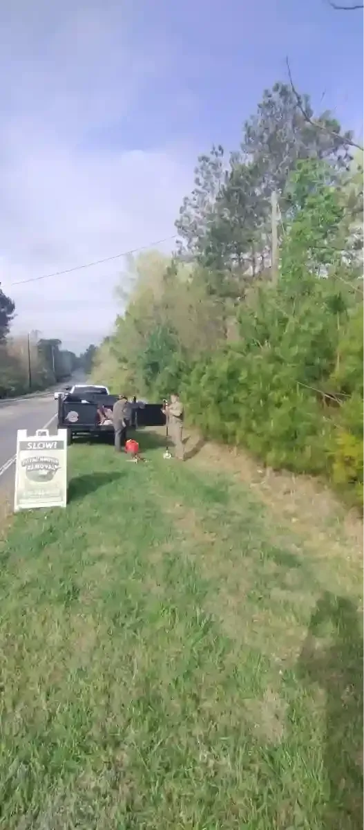 Two people stand beside a dark pickup truck parked on the grassy shoulder of a road near a woods edge.