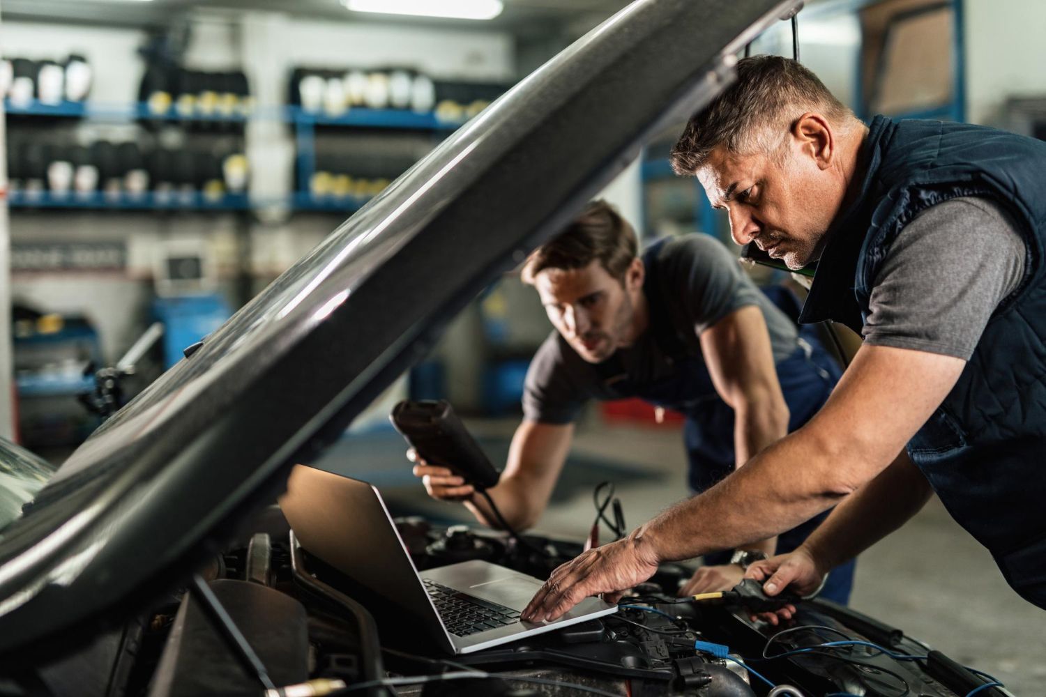 Dos mecánicos inspeccionando el motor de un coche en un taller, uno de ellos usando una tableta y con el capó abierto.