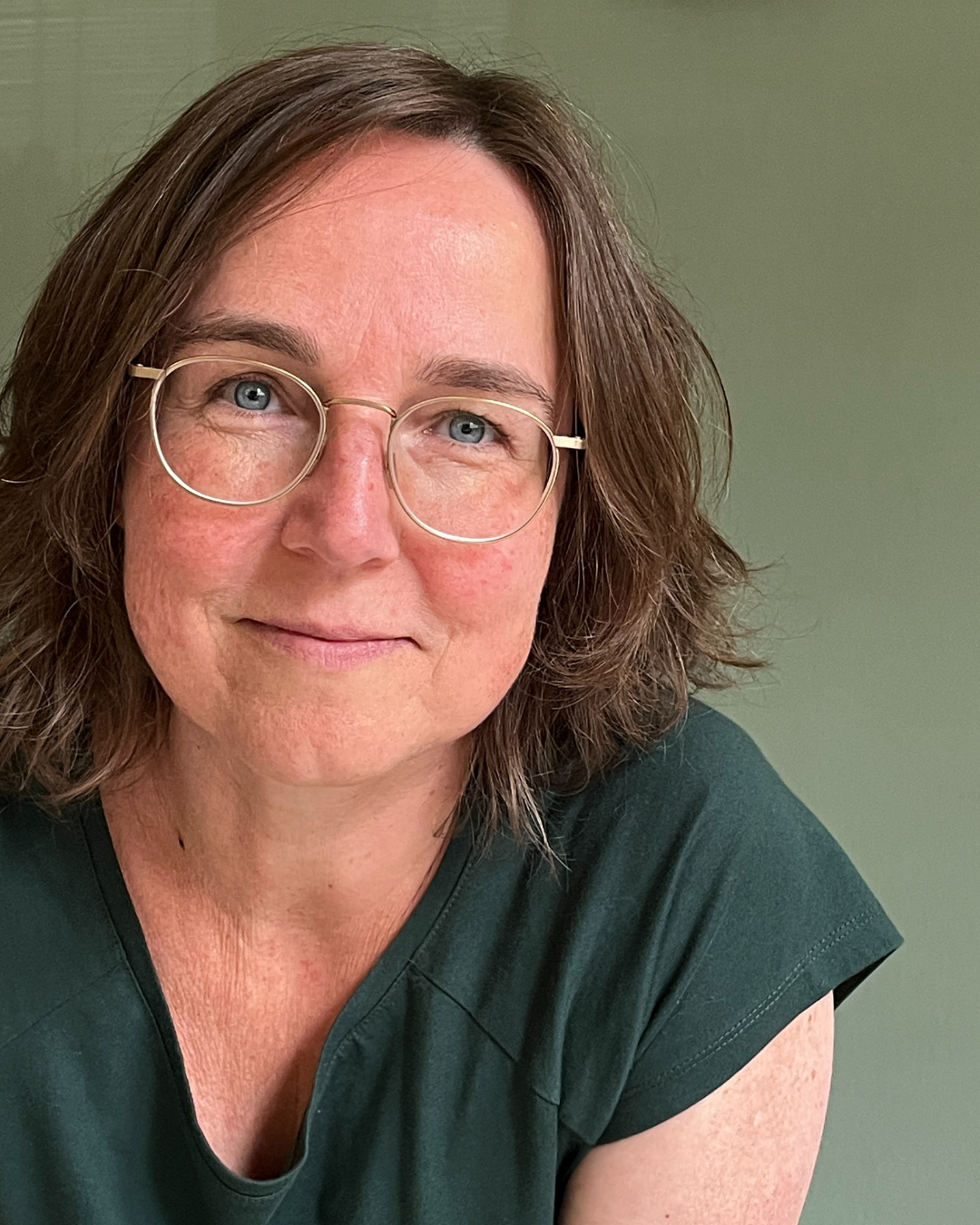 Astrid, de docent van de online yogaschool, zit op een yogamat. Ze is een vrouw met een bril en draagt een witte trui.
