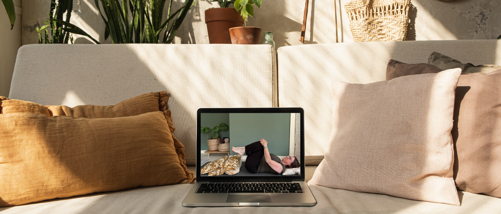 Laptop op de bank met daarop een yogavideo, omringd door kussens en planten.