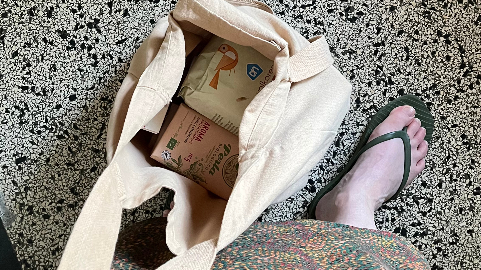 Beige tas met boodschappen, een persoon gekleed in groene teenslippers en een rok met print, op de gespikkelde vloer.