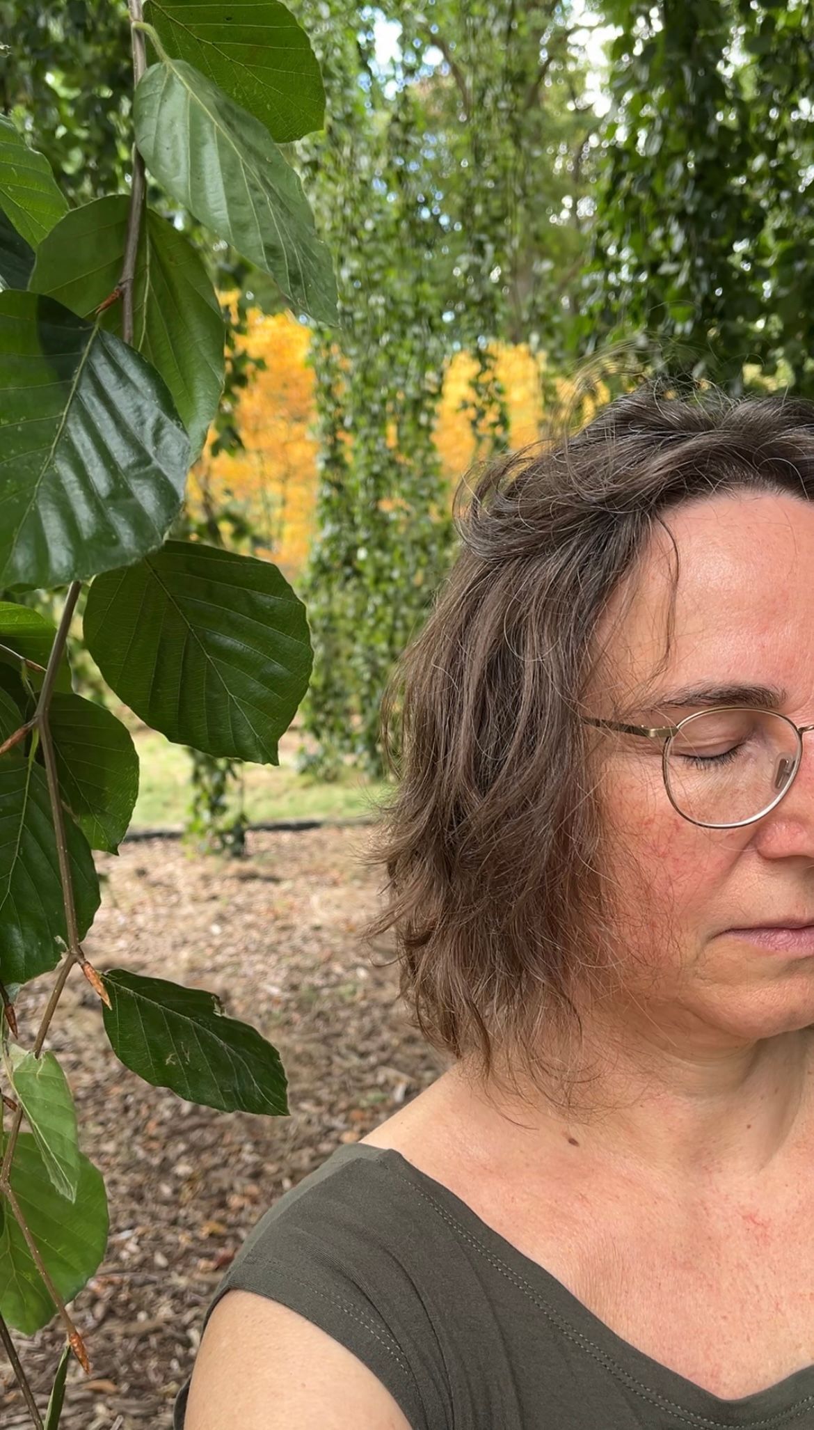 Vrouw met een bril en krullend bruin haar, in een parkachtige omgeving met wazig herfstgebladerte.