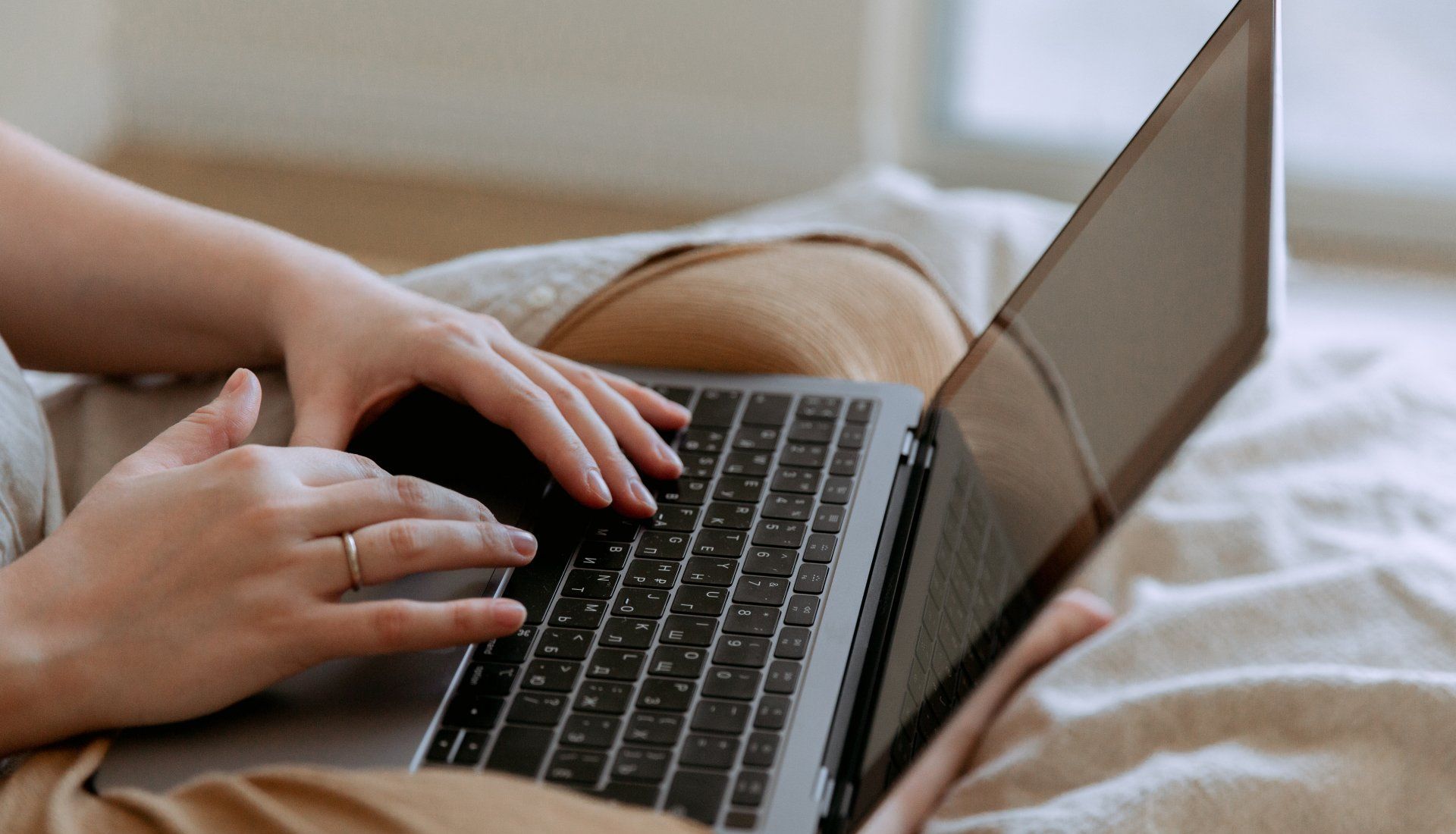 Persoon die met beide handen op een laptop zit te typen, zittend op een bed.
