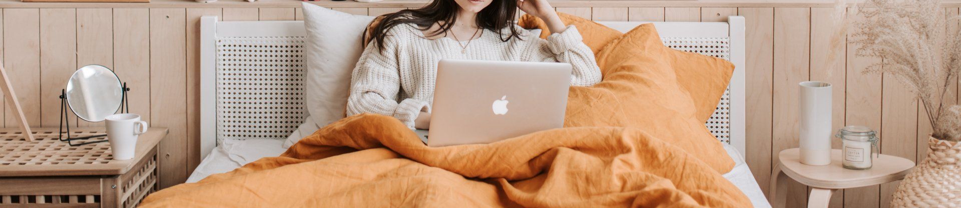 Persoon op bed met laptop, oranje deken en houten hoofdeinde.