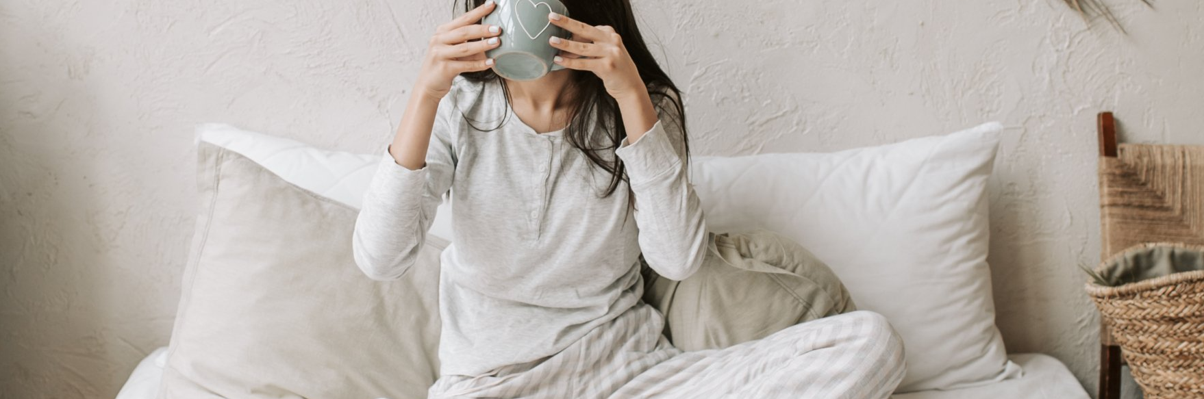 Vrouw in pyjama op bed, drinkend uit een mok. Lichtgekleurd beddengoed en muur.