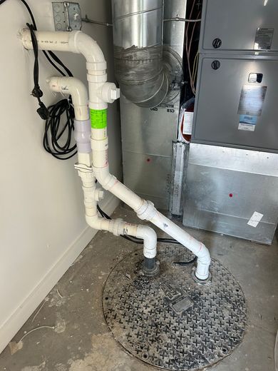 White PVC pipes connected to a circular sump pump basin on a concrete floor next to a metallic furnace unit.
