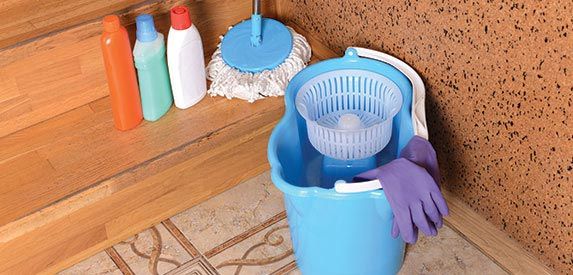 A blue bucket with a mop , gloves and cleaning supplies on the floor.