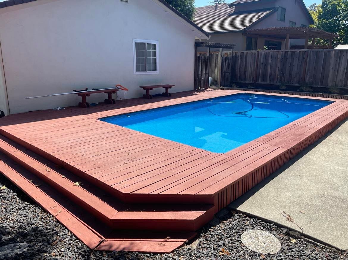 Small, rectangular backyard pool with surrounding red wooden deck and steps. Blue water and a white house.