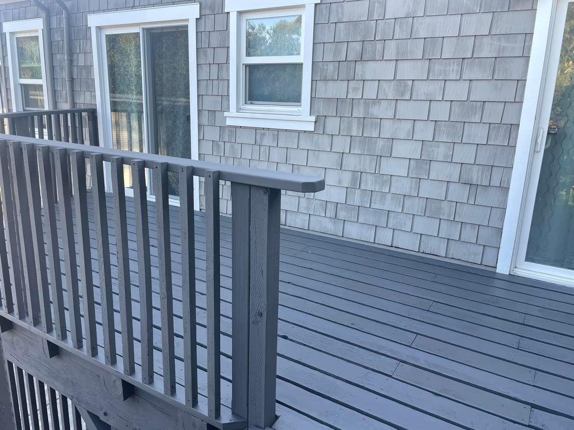 Gray wooden deck with railing, connected to a house with gray shingle siding and white-trimmed windows.
