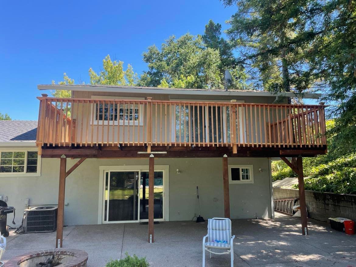 Wooden deck with railings attached to a gray house, supported by wooden posts. Sunny day, backyard.