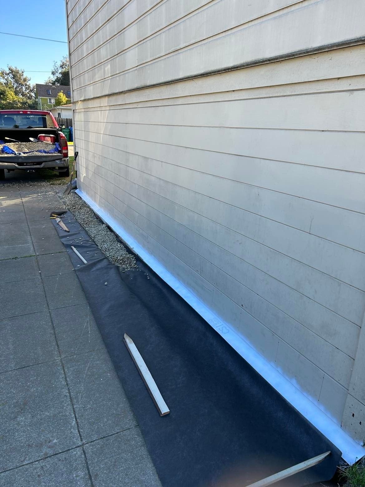 Exterior building wall with white siding, gravel bed, black fabric, and concrete sidewalk.