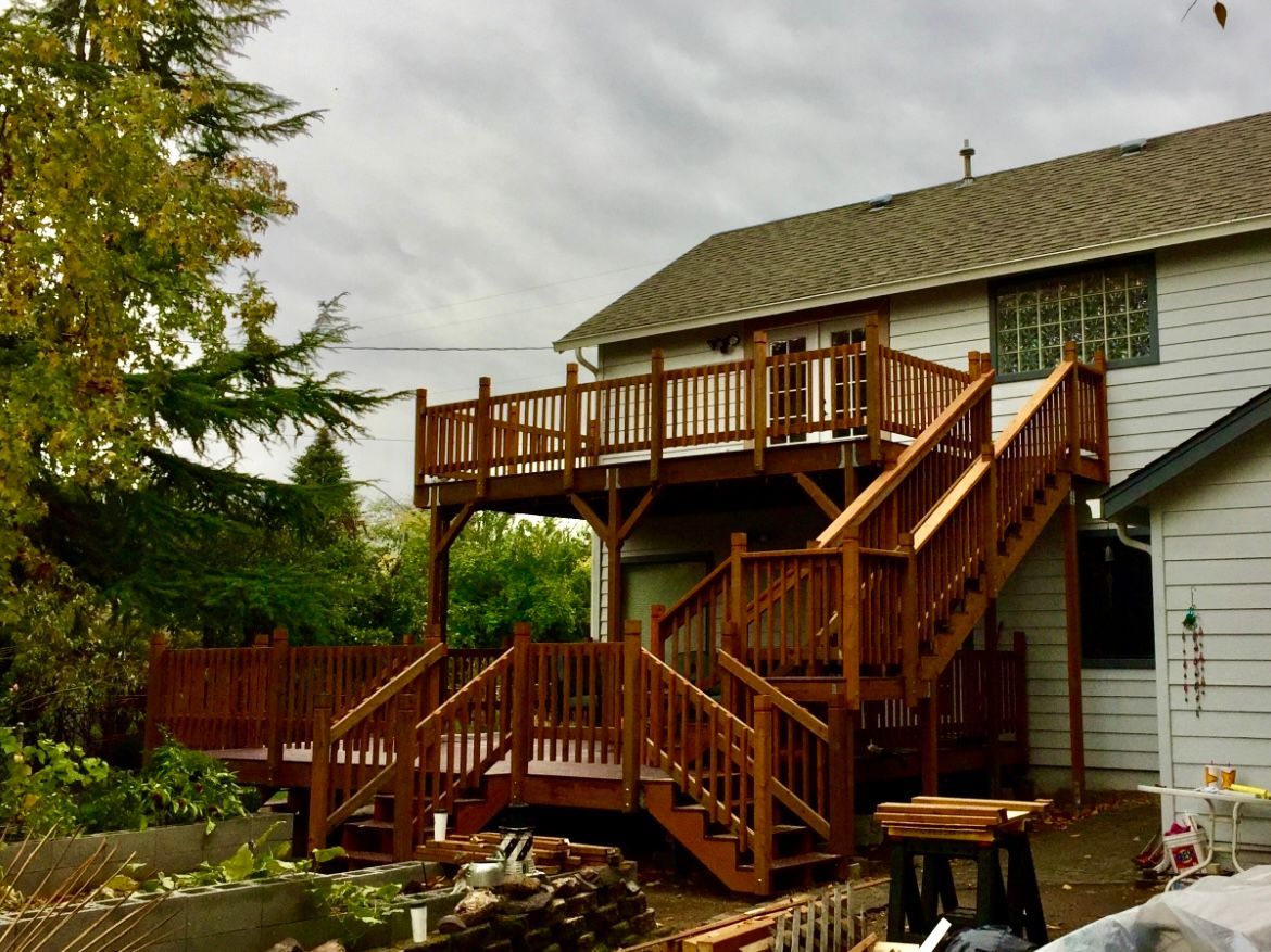 Two-level wooden deck attached to a house with stairs. Brown stained wood, overcast sky.