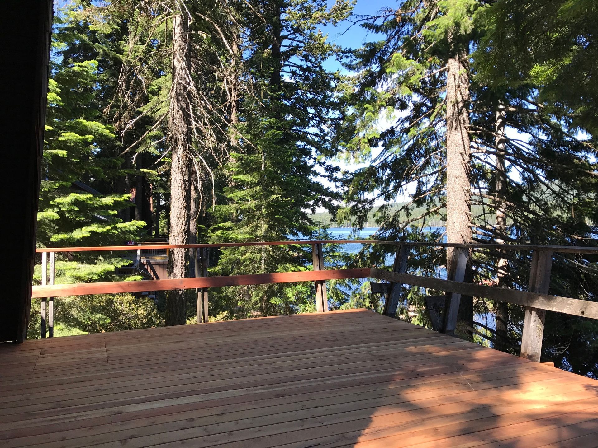 Wooden deck overlooking a lake, framed by trees and a wooden railing. Sunny day.