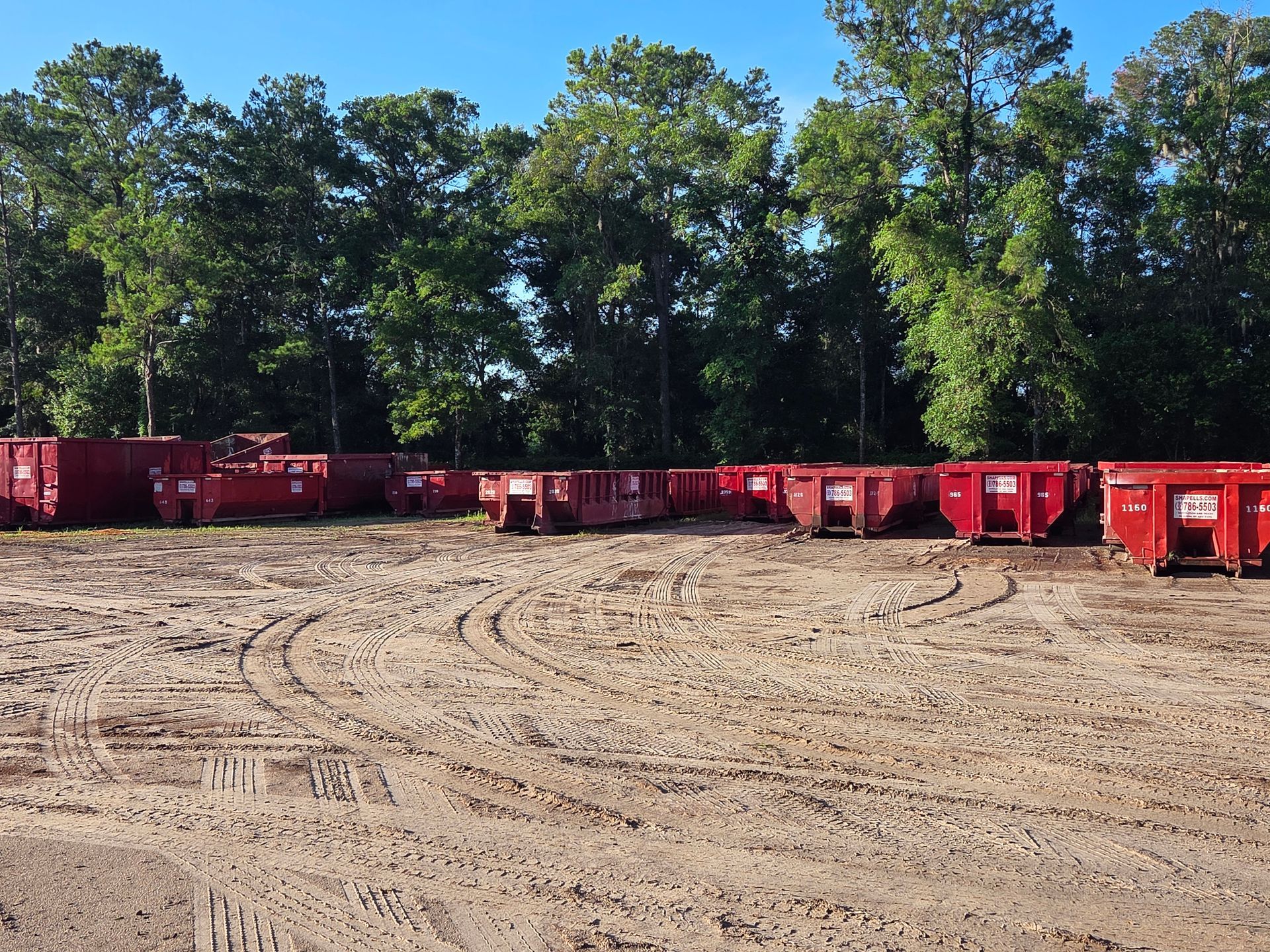 A row of red dumpsters in a dirt field with trees in the background