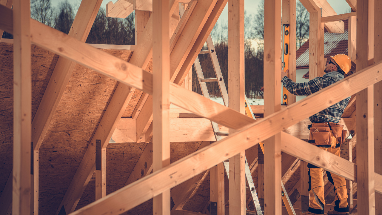 Carpenter using a level on wooden frame construction site. Yellow work clothes, hard hat.