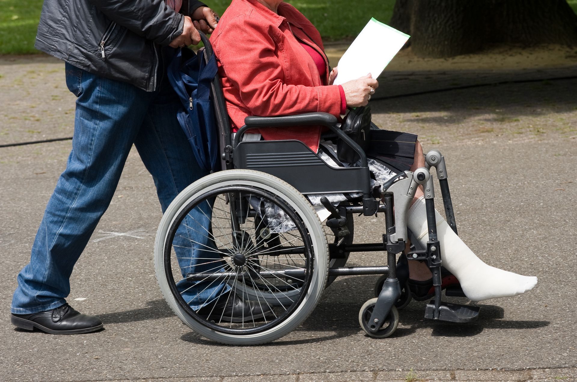 A person in a wheelchair covered with a beige blanket being pushed outdoors on a sunny day