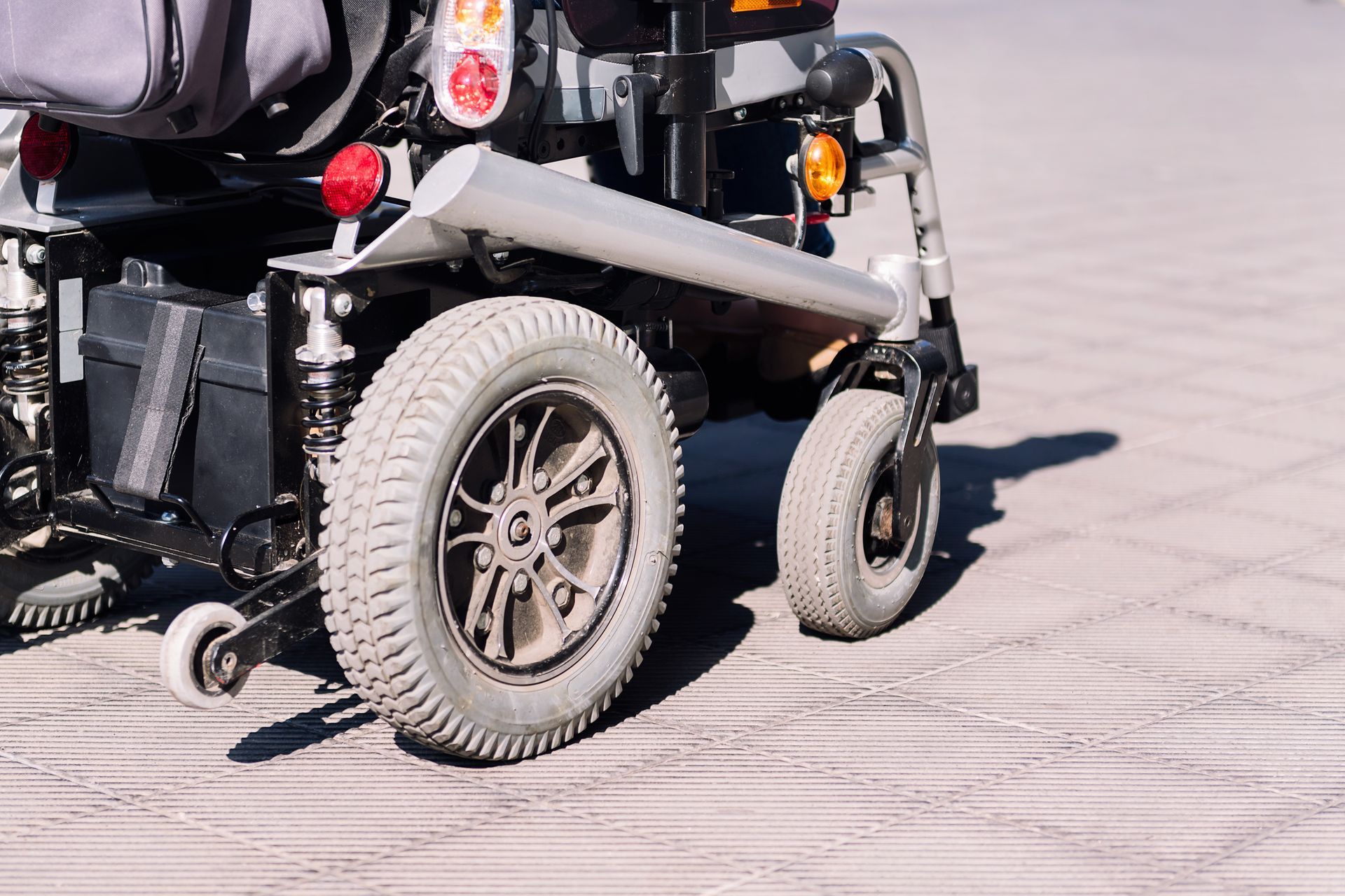 Rear view of an electric power wheelchair with large drive wheels parked on a sunny sidewalk. Rear view of an electric power wheelchair with large drive wheels parked on a sunny sidewalk.