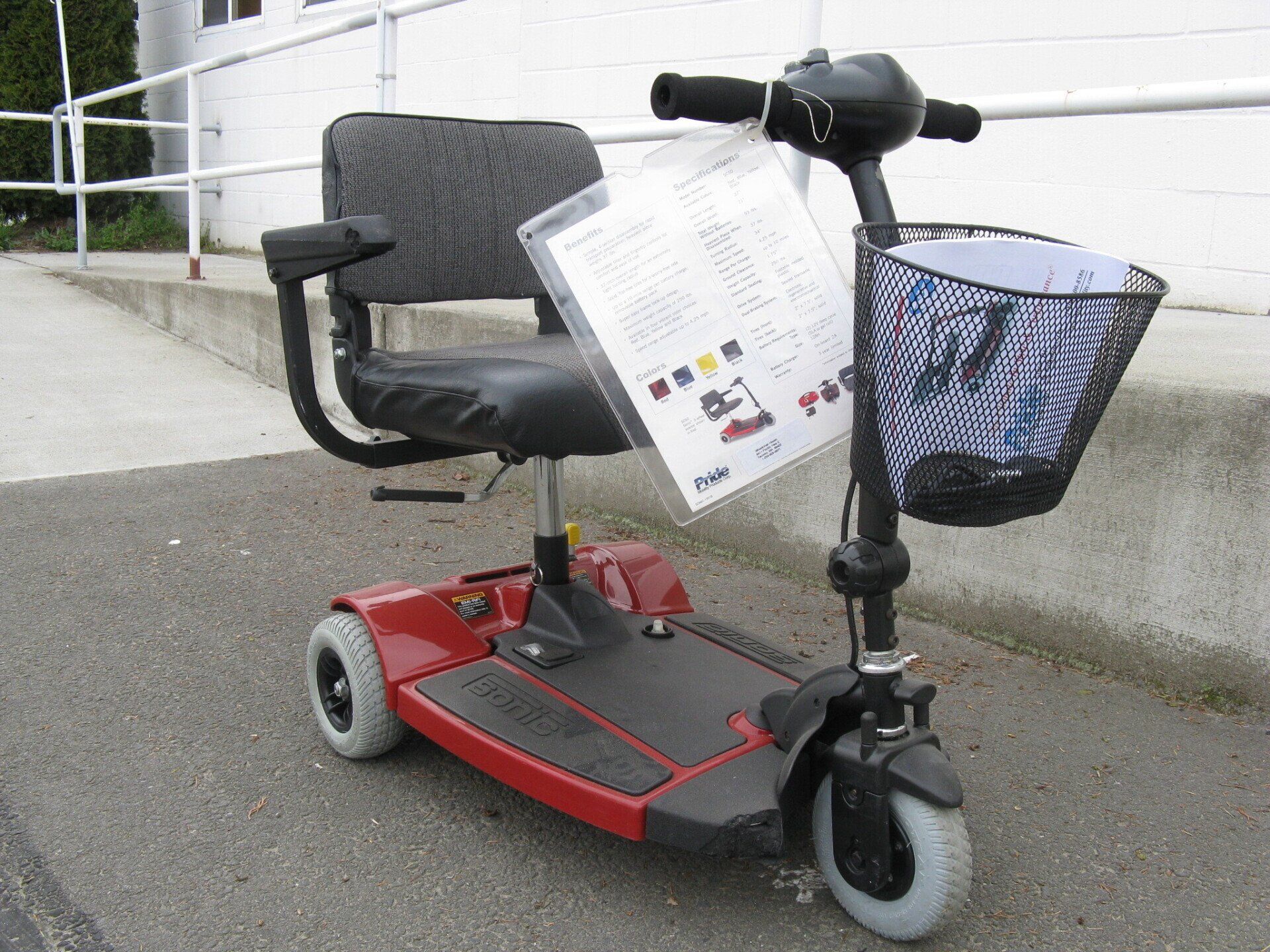 Red and black three-wheeled mobility scooter with basket and attached information sheet, parked outdoors.