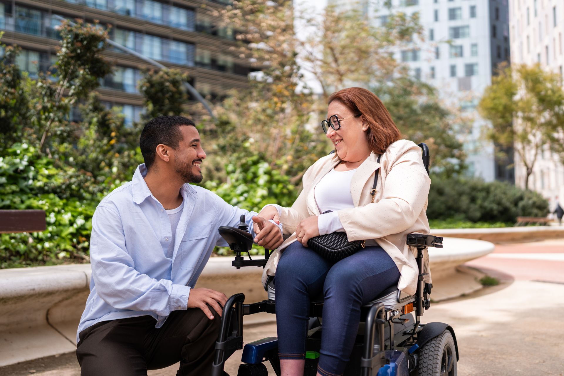 A young man holding hands with a smiling woman in a power wheelchair in a city park