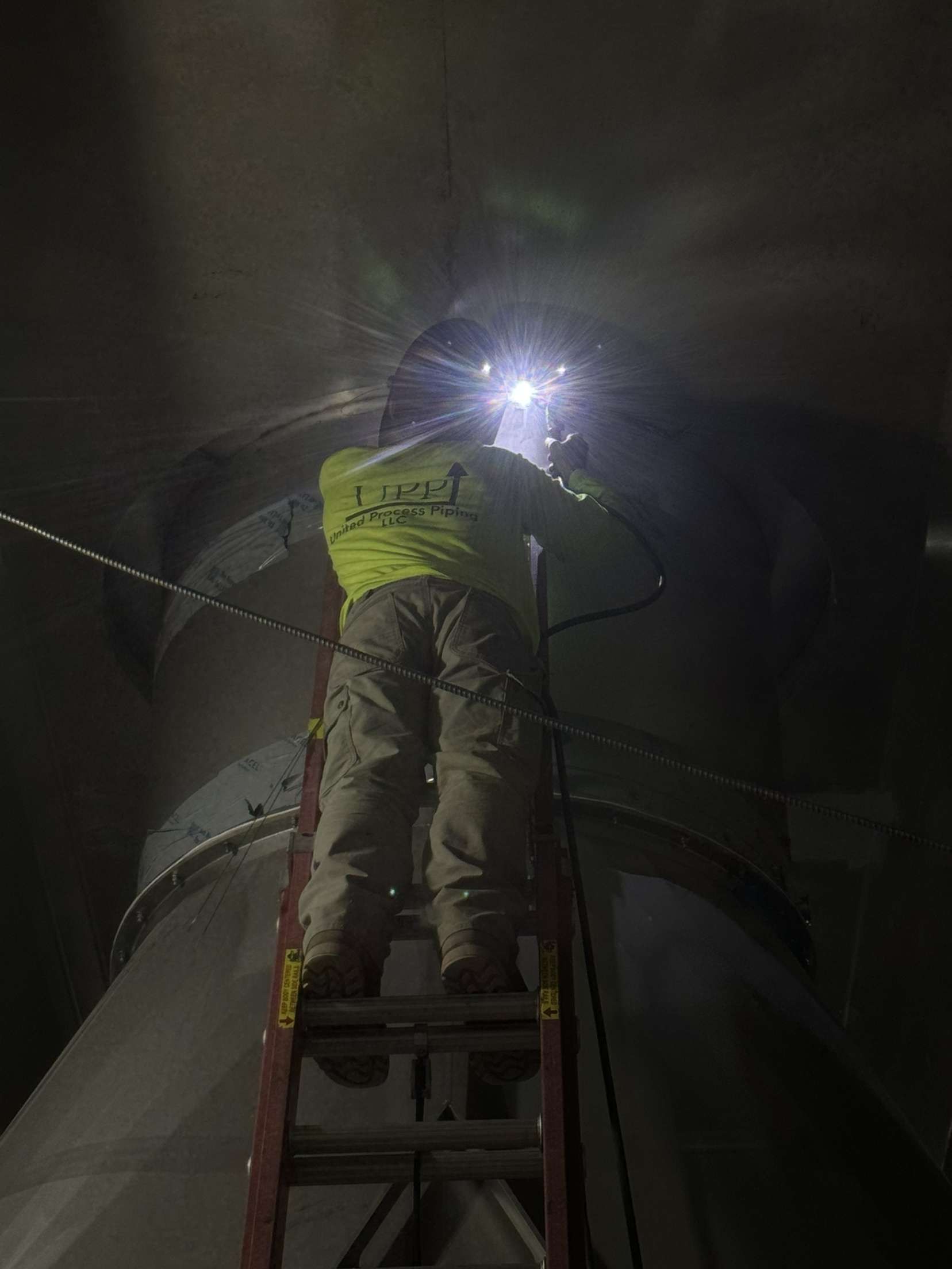 A man is standing on a ladder welding a pipe in a dark room.