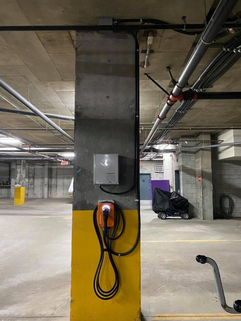 Une borne de recharge est suspendue à un poteau jaune dans un parking.