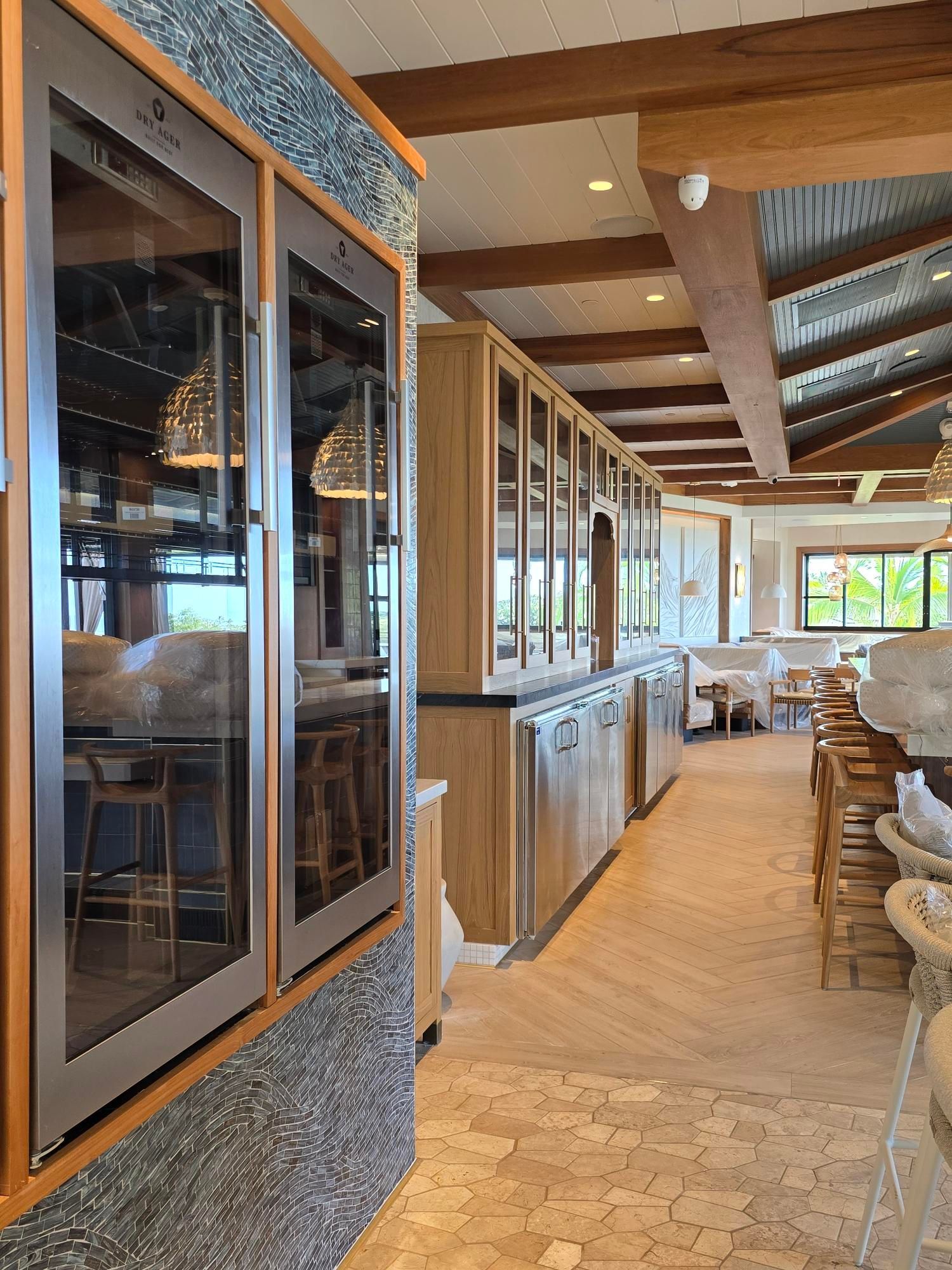 A restaurant interior with wooden cabinets, wine coolers, and a tiled floor. Dining tables and a bar are in the background.