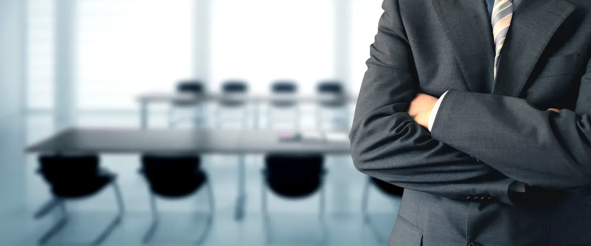 A man in a suit and tie is standing in front of a conference room with his arms crossed.