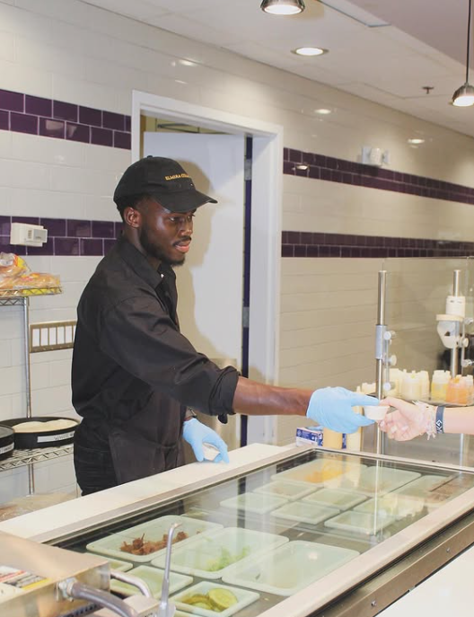 Man in a black uniform and cap serving food behind a deli counter, wearing gloves.
