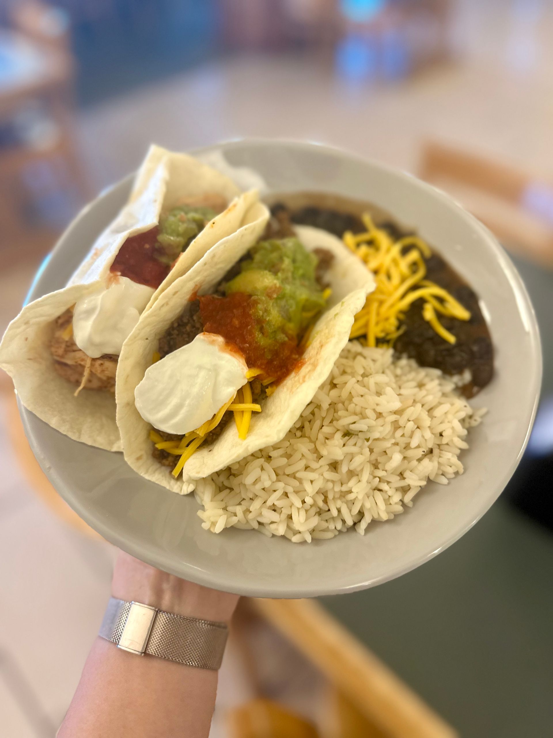 Plate of tacos with guacamole, sour cream, cheese, rice, and beans, held by a person.