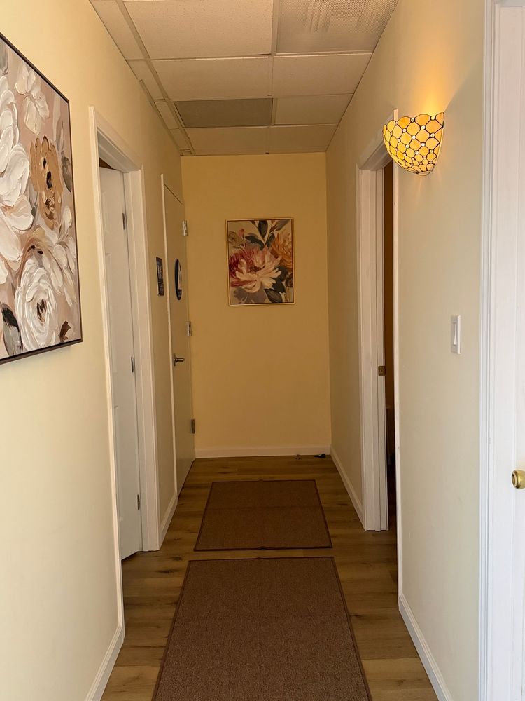 Small treatment room with a single bed, chair, side table, and wall art in soft beige tones