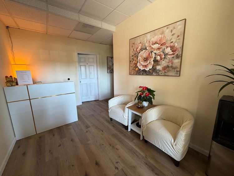Small treatment room with a single bed, chair, side table, and wall art in soft beige tones