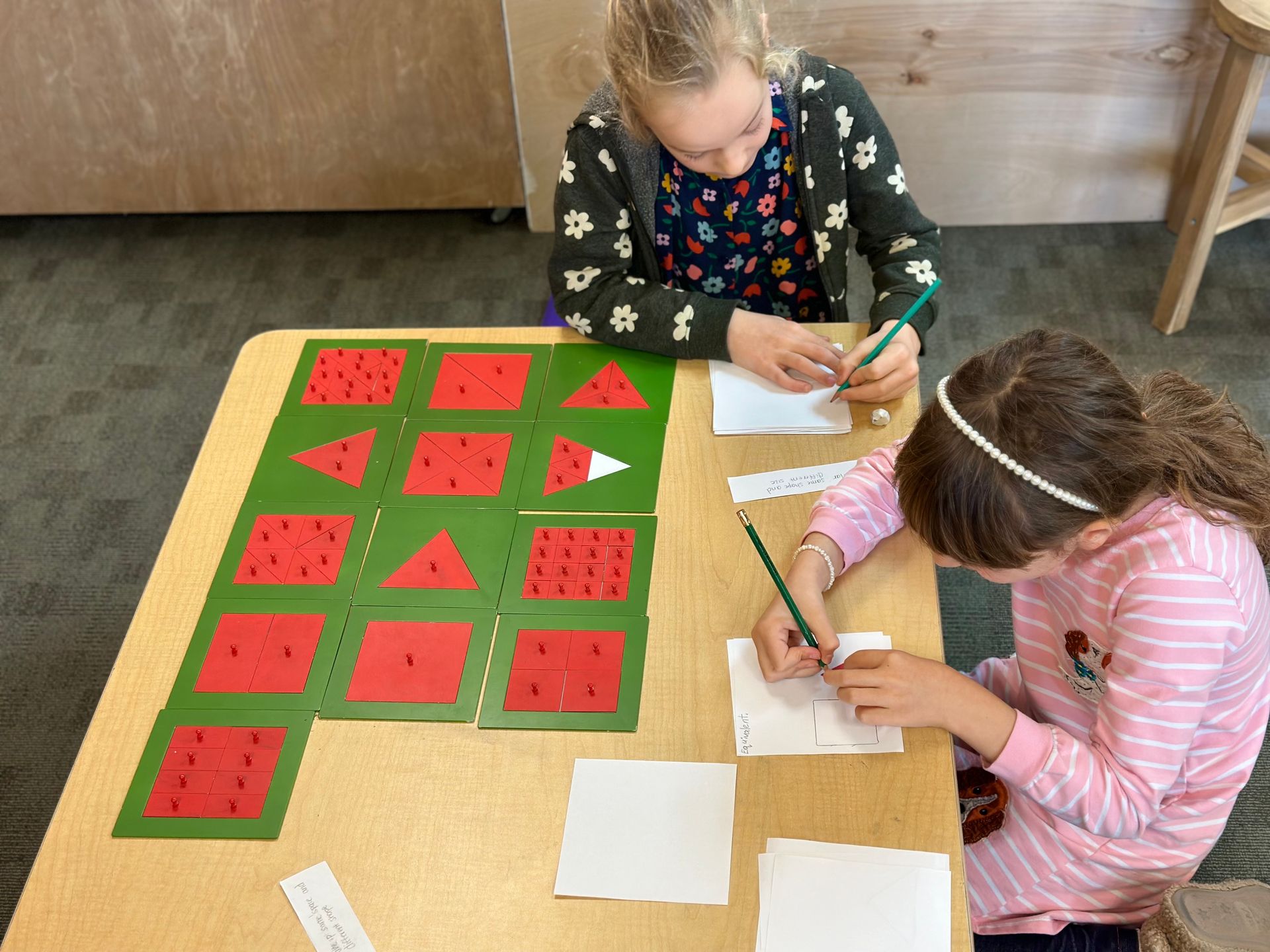 Two Lower Elementary students working together on a Montessori math material called Congruency, Similarity, and Equivalents with metal insets. This material also helps to understand fractions.