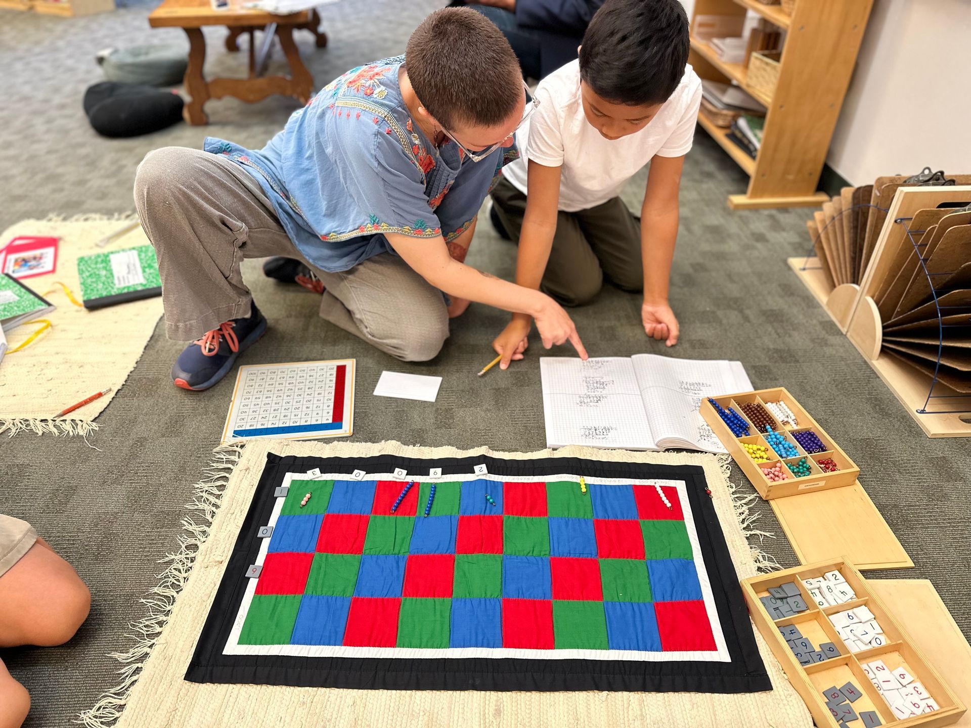 An Upper Elementary teacher works with a student on a Montessori math material called the Checkerboard. Checkerboard is for long form multiplication.