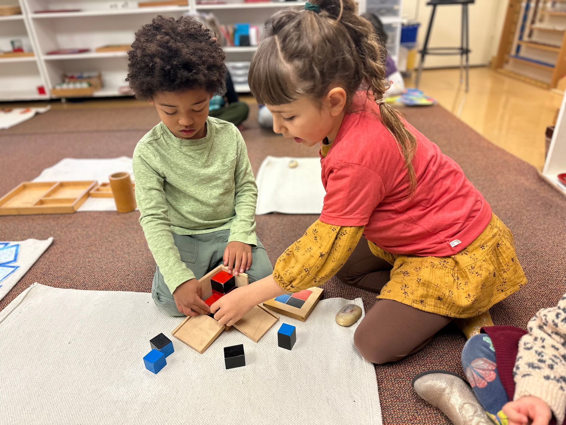 One older Kindergarten Bridge student helping a younger student in the classroom with the Montessori Binomial Cube.