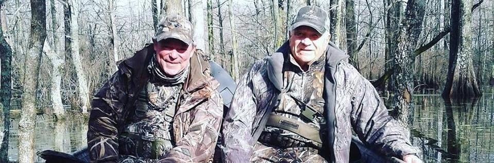 Two hunters in camouflage sitting in a wooded area, smiling at the camera.