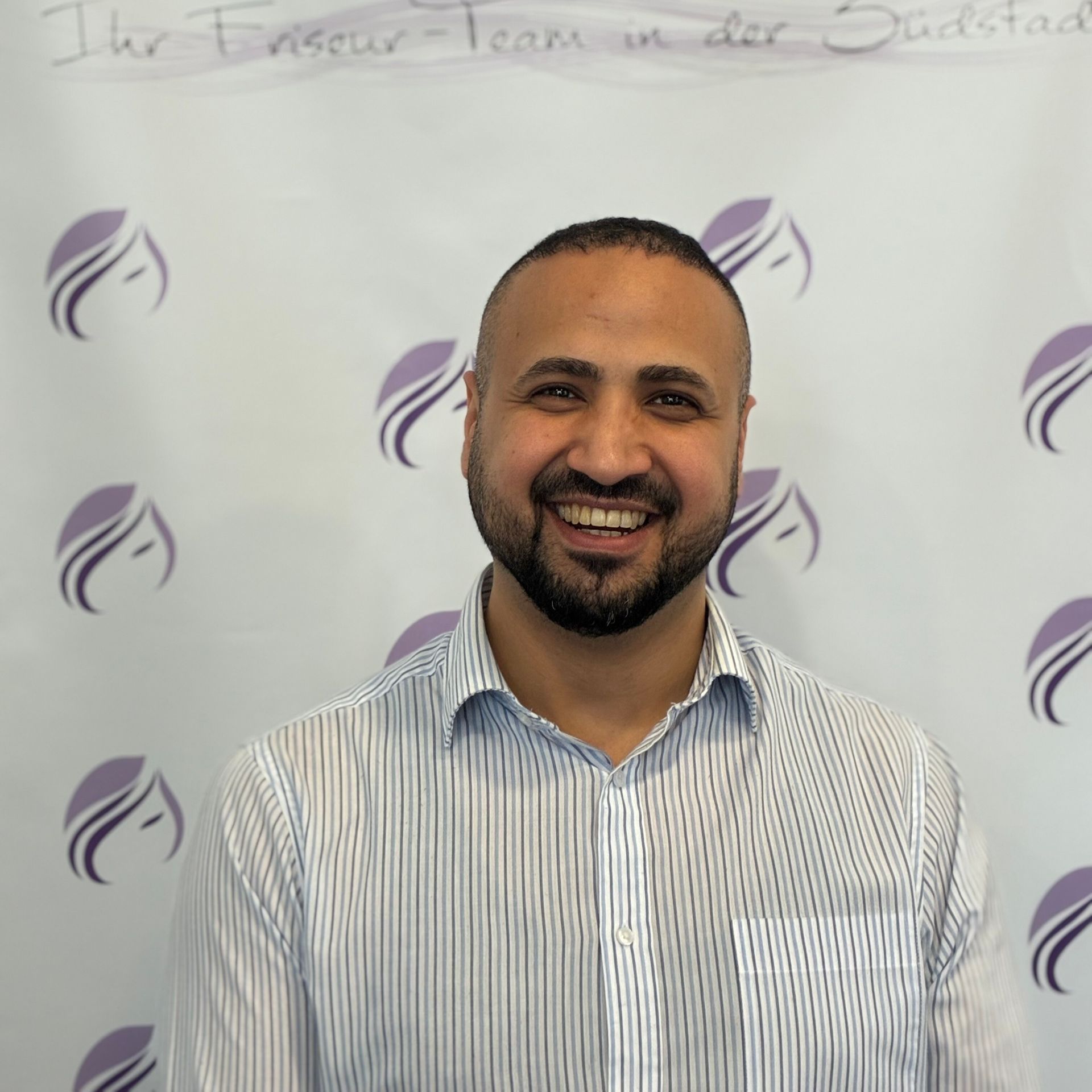 Ein Mann mit Bart lächelt vor einer Wand mit lila Logos darauf