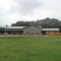 Single-story gray house with a covered porch, set behind a large green lawn under cloudy skies.