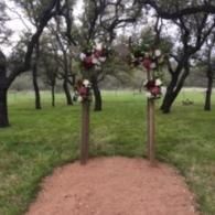 Outdoor archway decorated with flowers in a grassy grove, with a dirt path leading through it