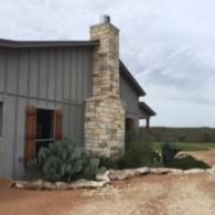 Ranch-style house exterior with stone chimney, gray siding, and desert landscaping on a dirt drive
