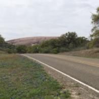 Empty rural road curving past dry hills and sparse trees under a cloudy sky