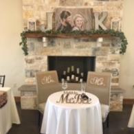 Wedding reception table with white cloth, wooden initials, and a fireplace mantel decorated with greenery and photos